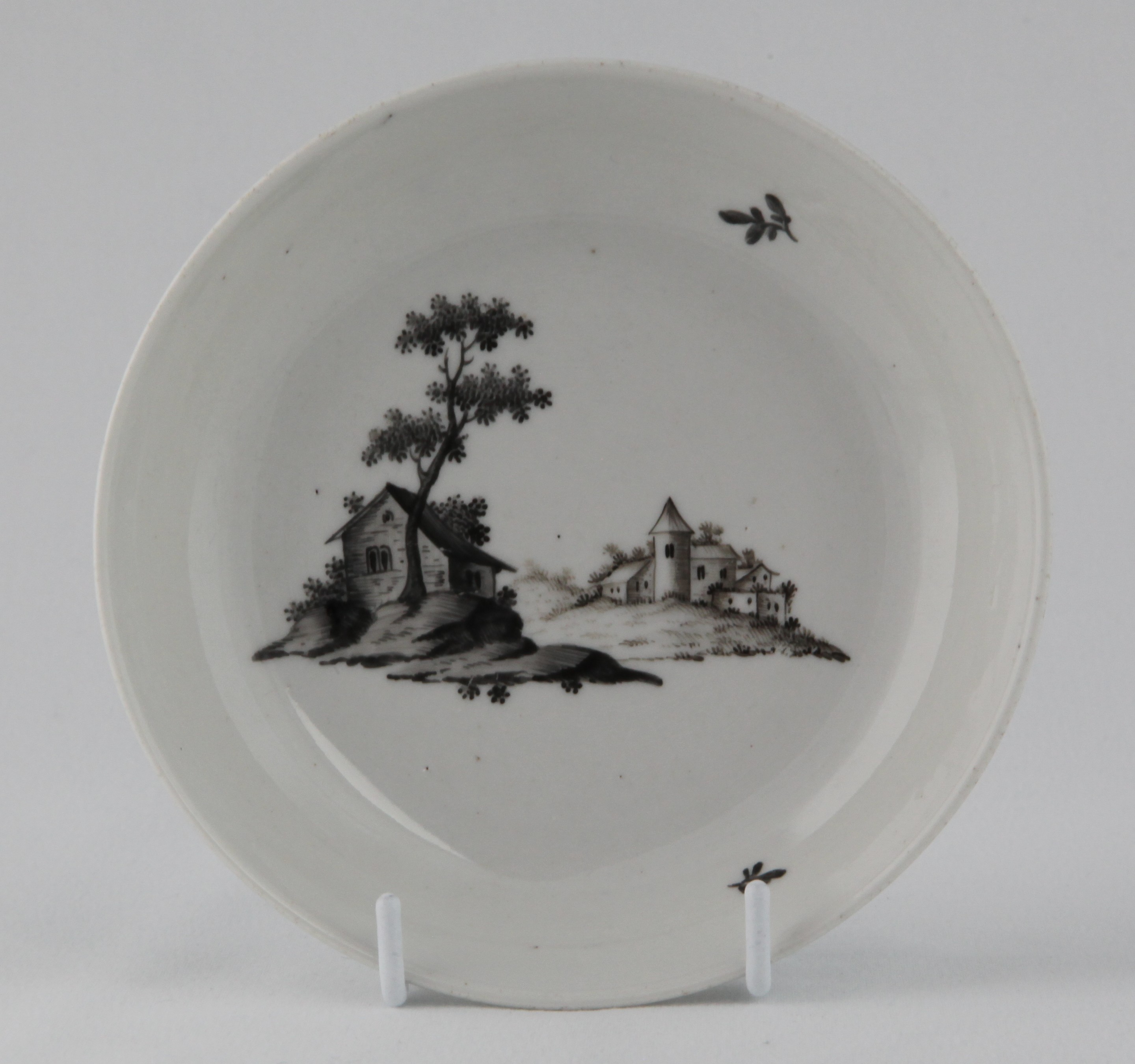 A rare Bristol saucer, unusually painted en grisaille with buildings in a European river landscape, and two leaf sprigs, 5 1/4" diameter, circa 1775, faint blue painted X mark