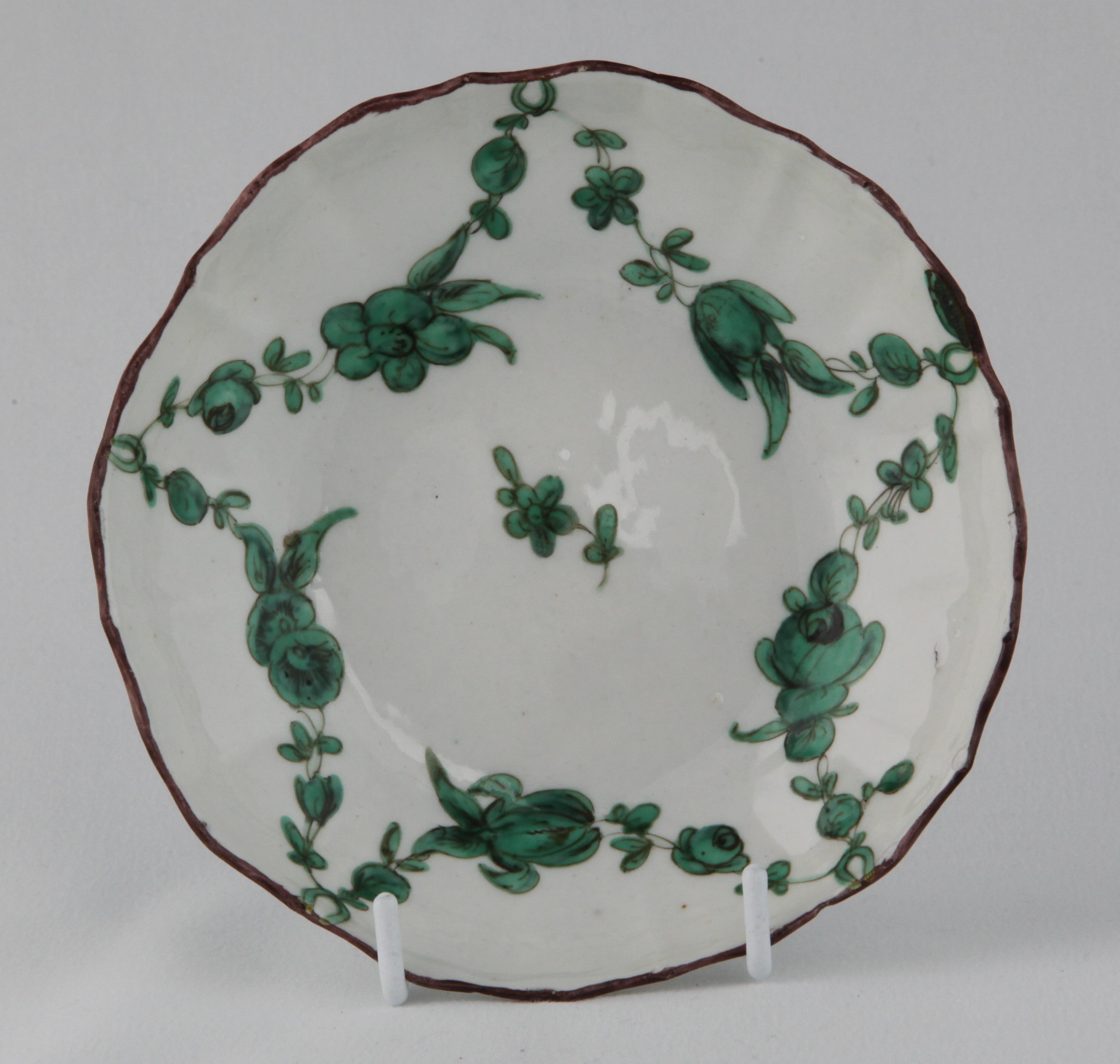 A Bristol ogee shaped coffee saucer, painted in green and black with floral garlands, suspended from rings, within brown line borders, circa 1775, blue painted B7 mark