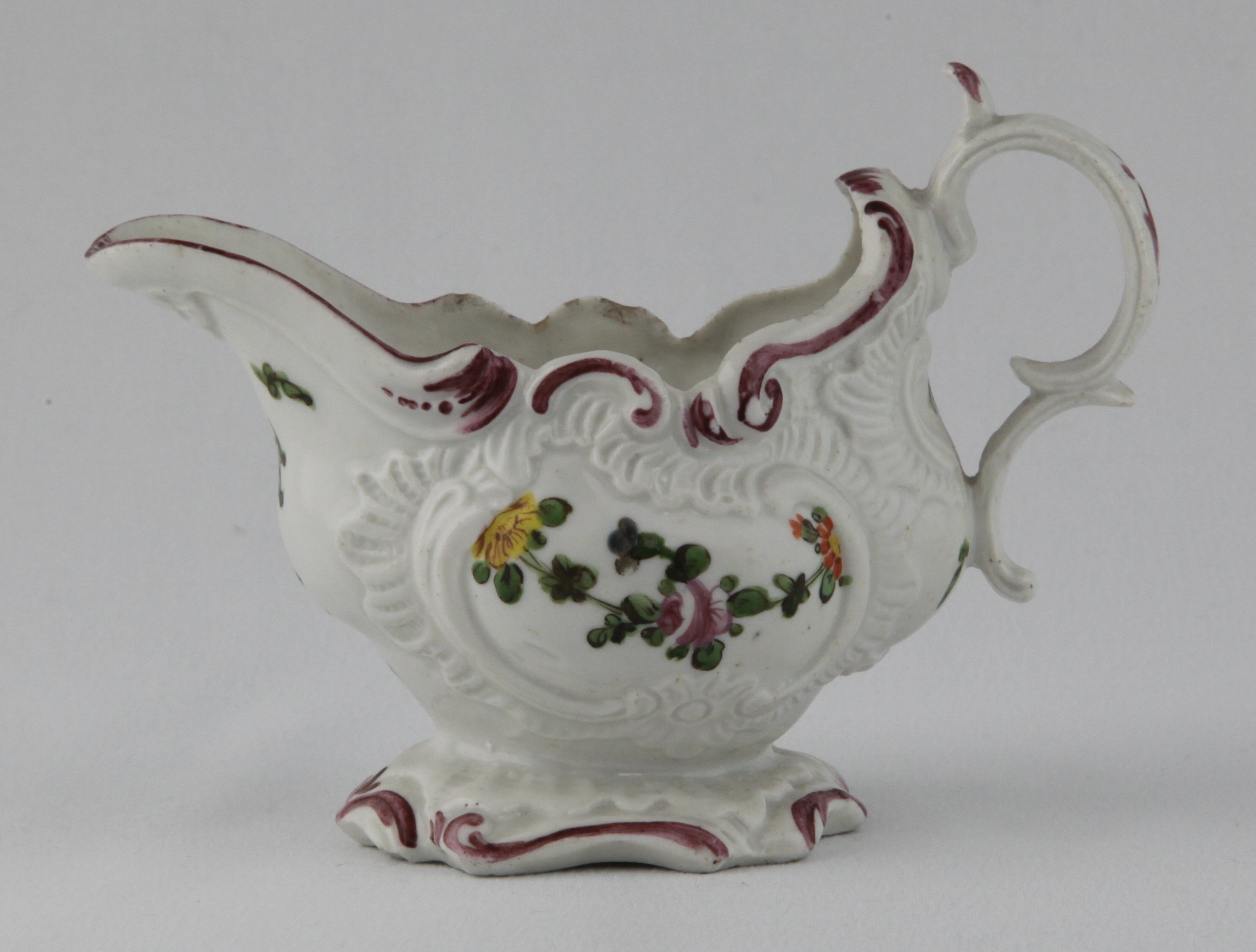 A Plymouth oval cream boat, of rococo silver form, with double 'C' scroll handle, painted in Chinese famille verte palette with flowers and leaves, within shell and scroll moulded cartouches, and with scattered flowers and fruits, the shell and scroll moulded spreading foot with a puce band, the interior painted with a leaf spray, circa 1770, no mark