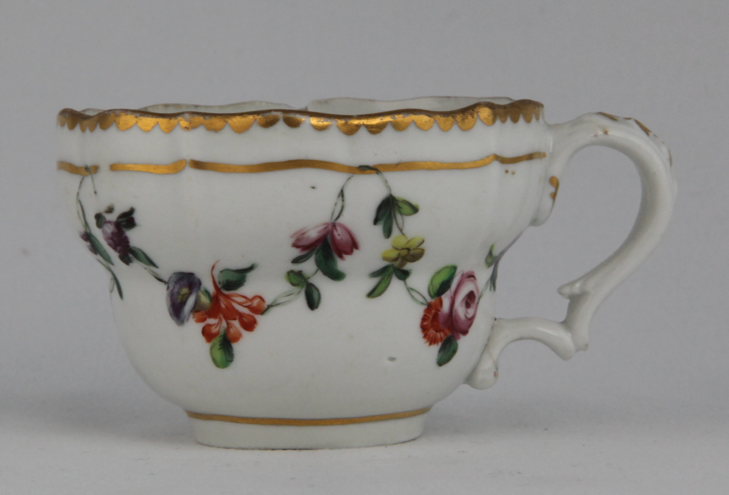 A Bristol porcelain teacup of ogee shape and harebell moulded C scroll handle, painted in enamels with garlands of flowers suspended from a gilt line, under a gilt dentil rim
