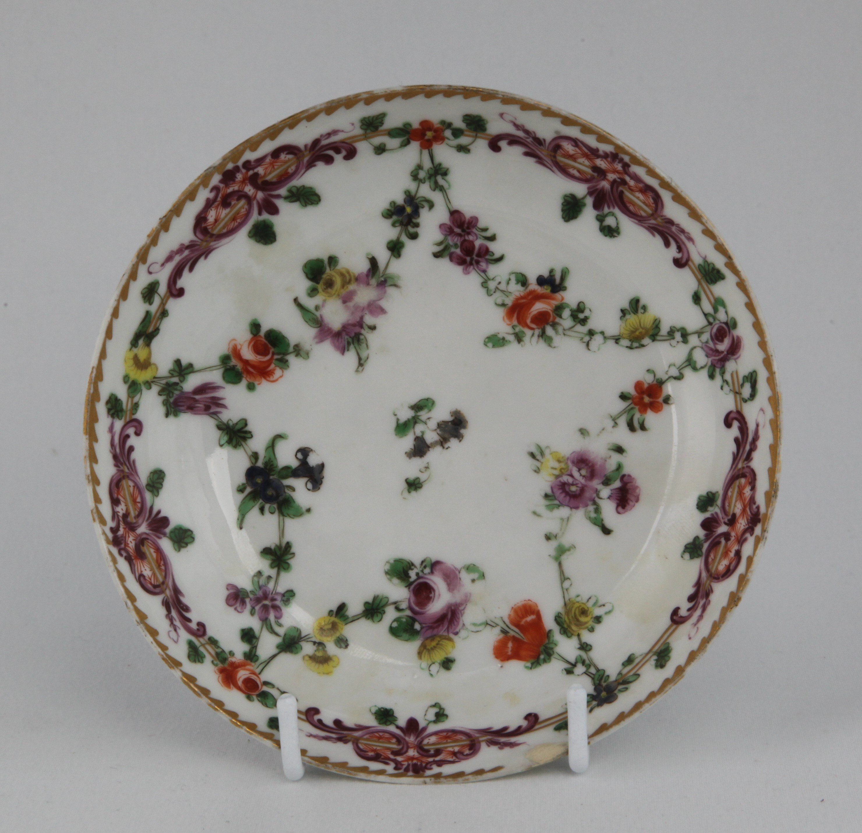 Saucer, decorated by No mark, A Bristol saucer, painted in enamels with garlands of flowers suspended from a double gilt line which supports magnificent puce and orange scroll cartouches under an angular gilt dentil rim. — Plymouth & Bristol porcelain piece 1684.1