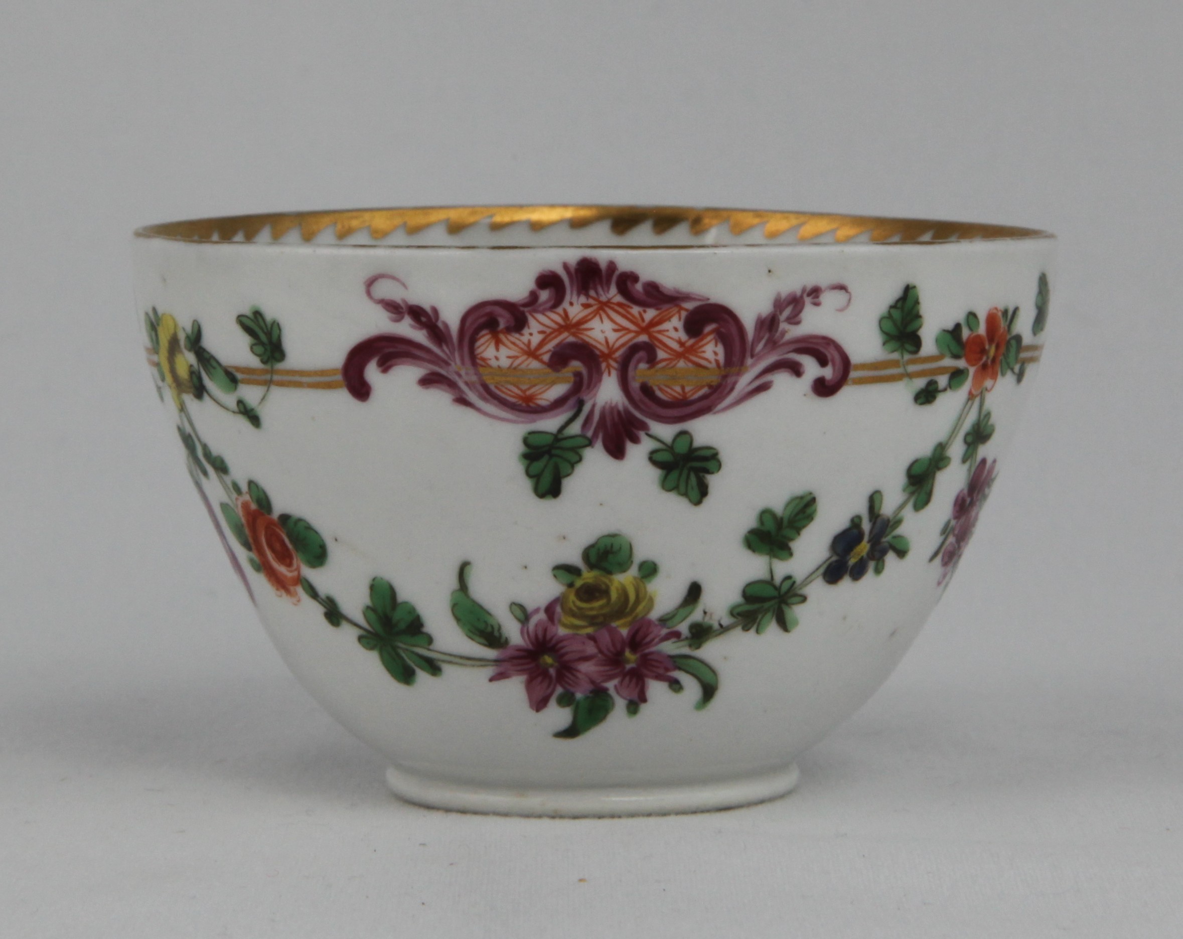 A Bristol teacup with elaborately moulded handle, painted in enamels with garlands of flowers suspended from a double gilt line which supports magnificent puce and orange scroll cartouches, with an angular gilt dentil rim to the inside.