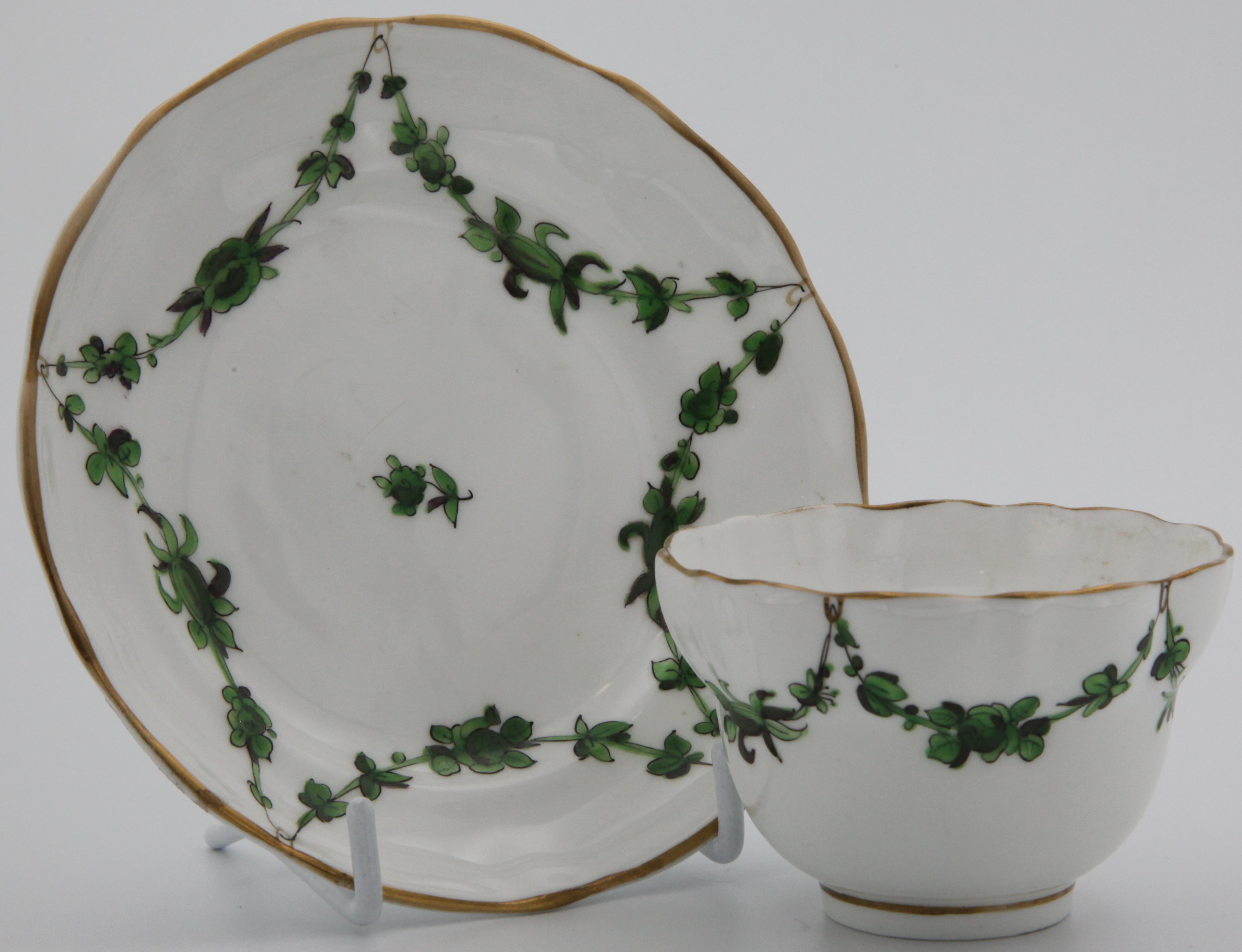 A Bristol-style replacement tea bowl and saucer with green monochrome floral garlands suspended from gilt loops.
