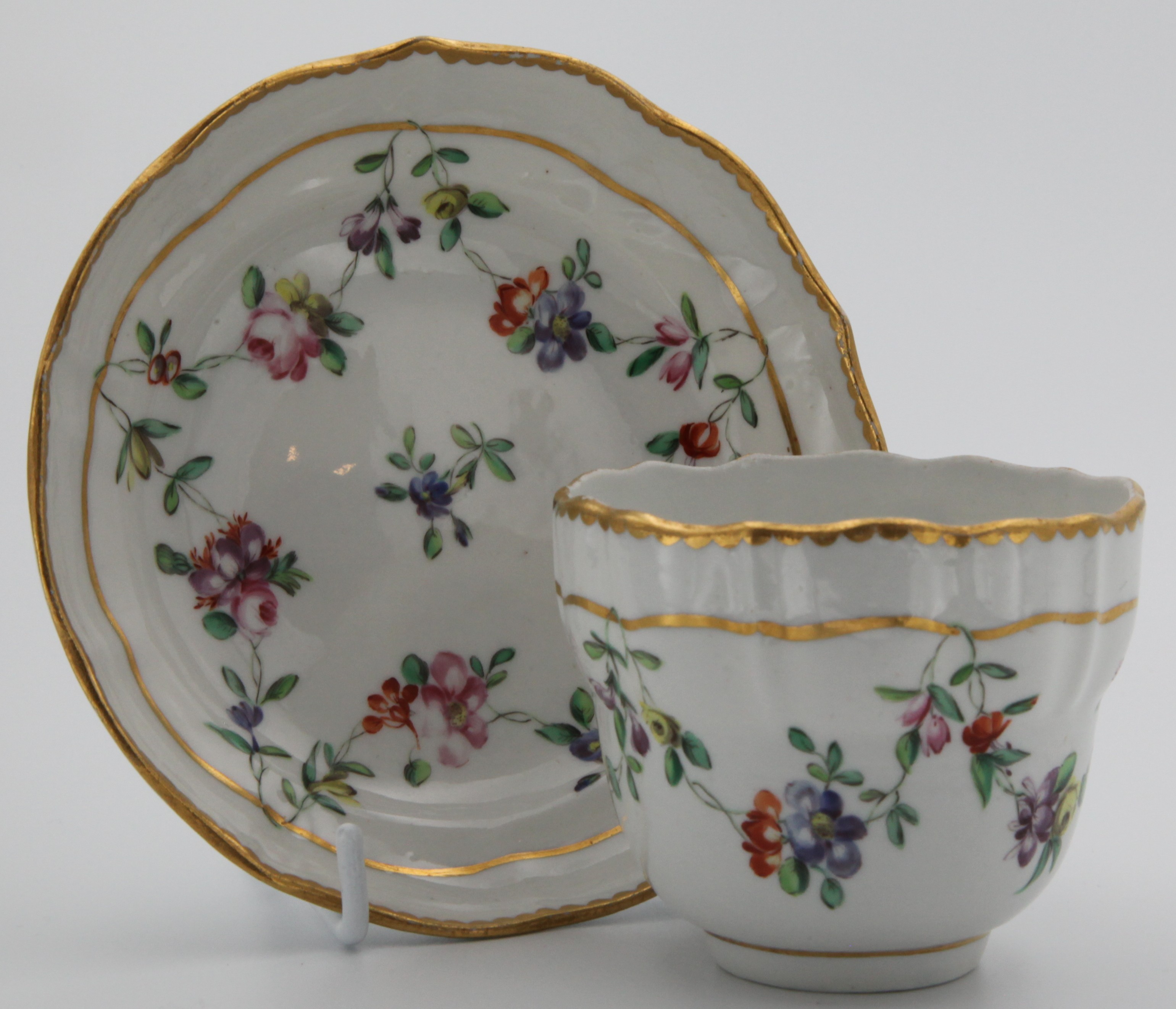 An ogee-shaped Bristol coffee cup and saucer with colourful garlands suspended from a gilt line and with gilt dentil rim.