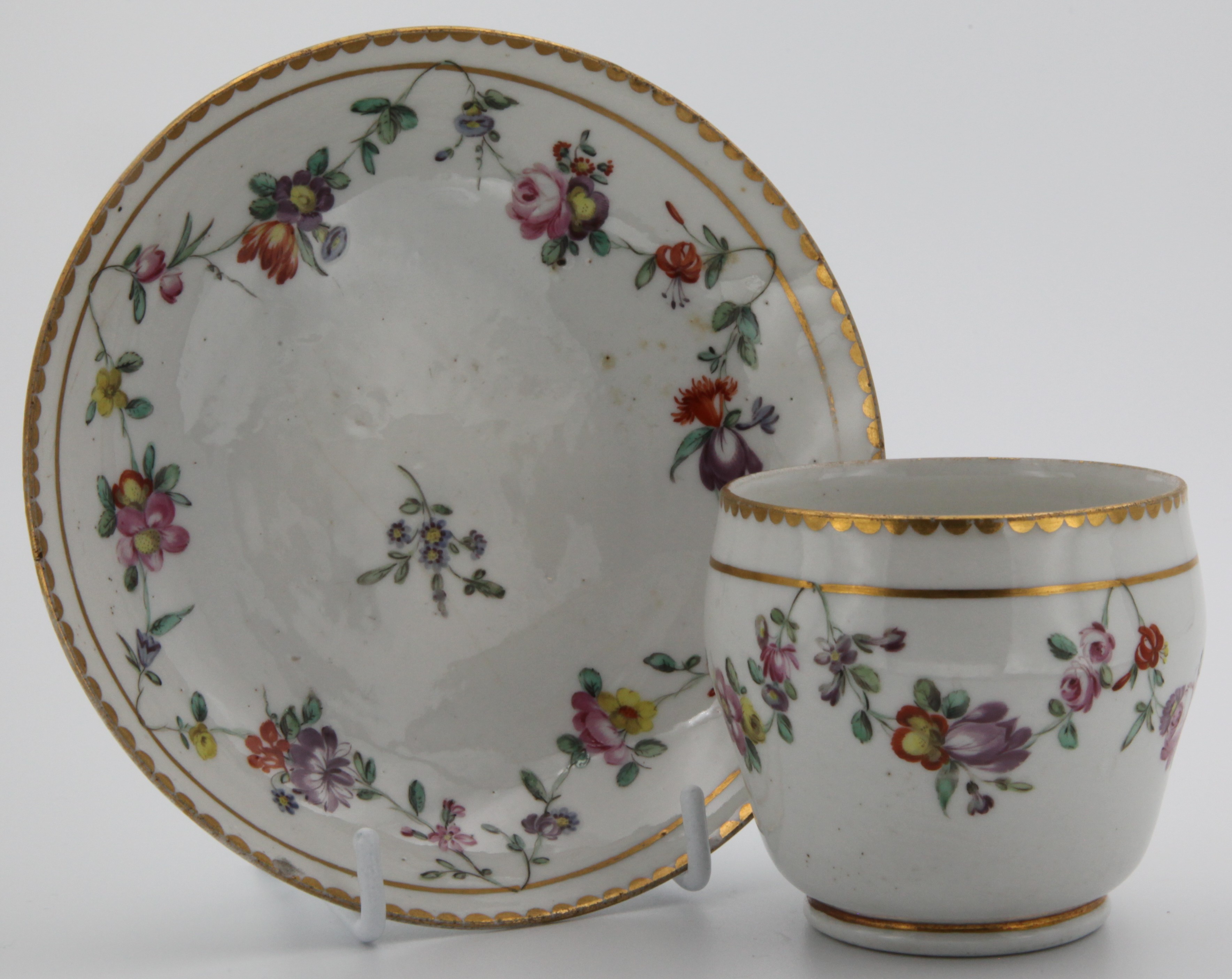 An ogee-shaped Bristol coffee cup and saucer with colourful garlands suspended from a gilt line and with gilt dentil rim.