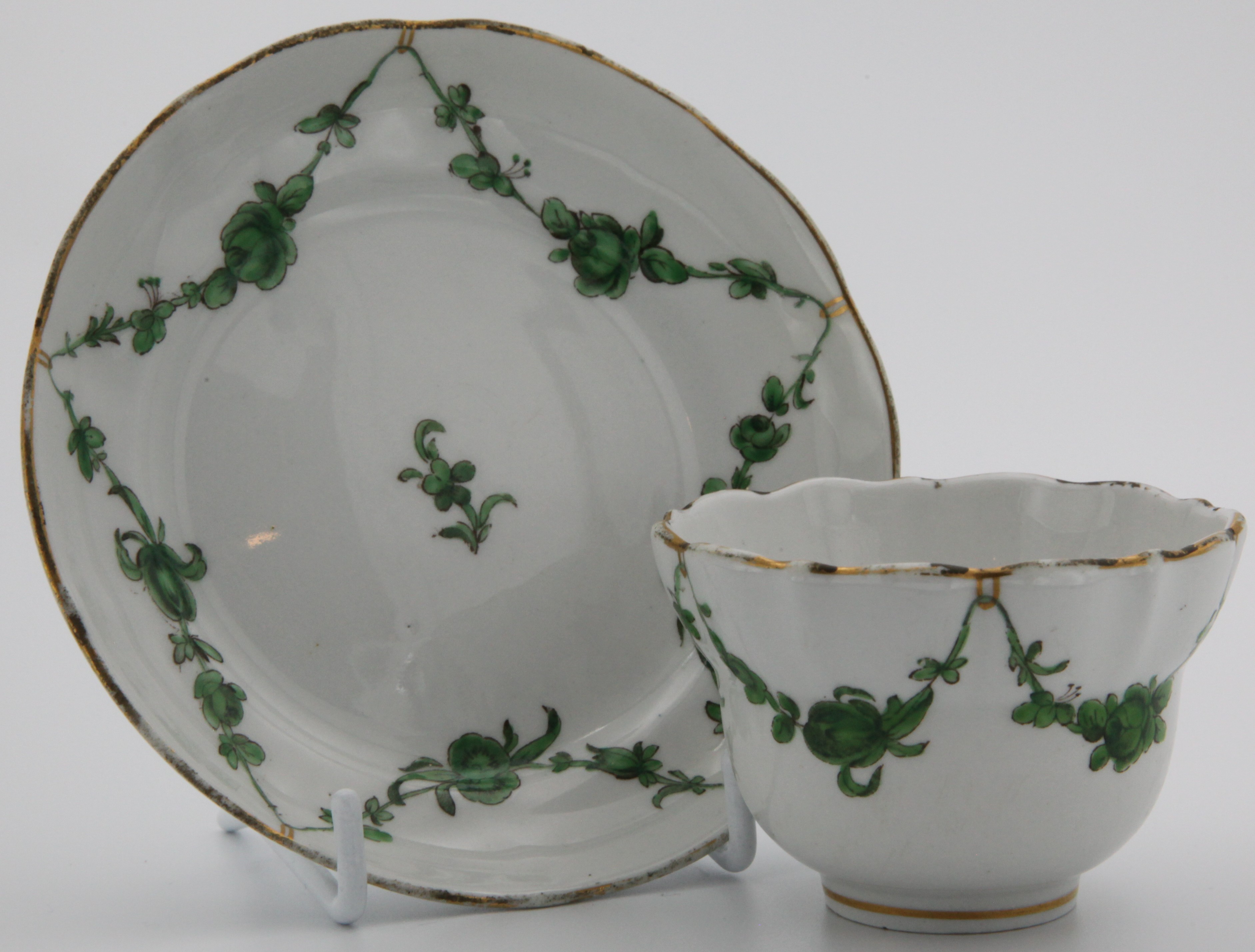 An ogee-shaped Bristol tea bowl and saucer with green garlands suspended from gilt loops hanging from gilt dentil rim.