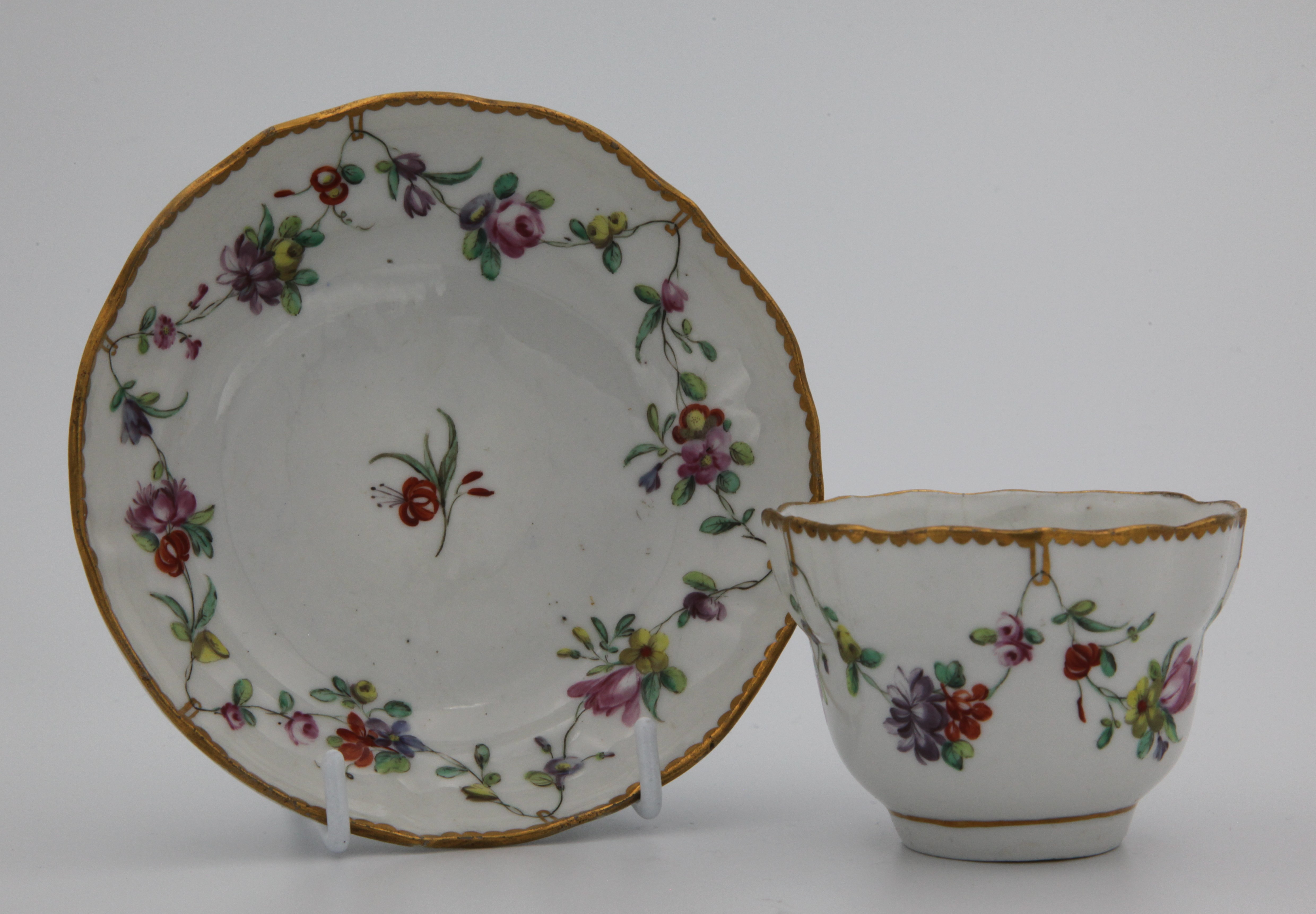 A Bristol tea bowl and saucer with garlands of flowers suspended from gilt loops hanging from gilt dentil rim.