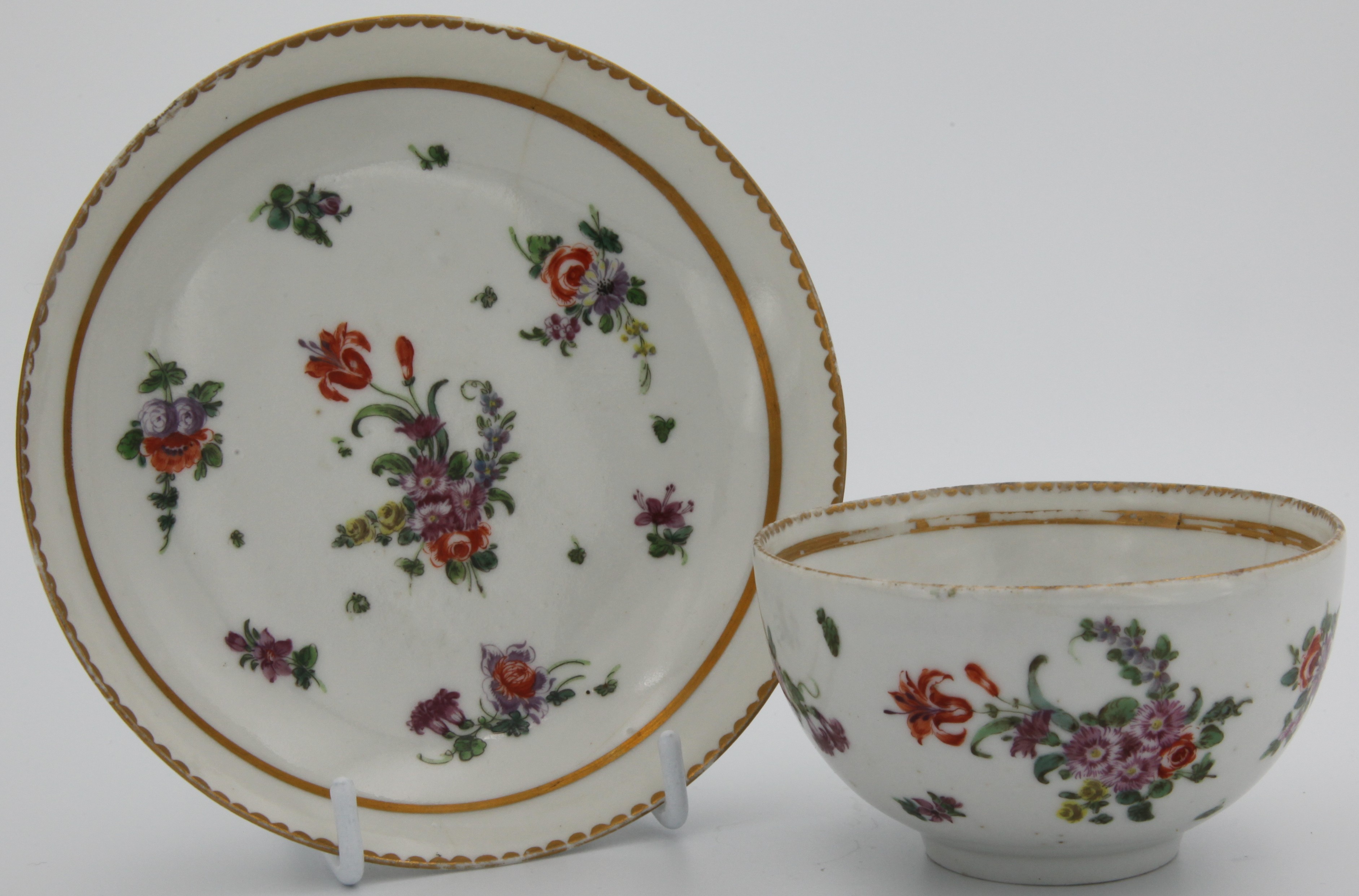 A Bristol cup and saucer with with a central spray featuring a prominent orange flower and scattered sprays and leaves inside a thick gilt band, surrounded by gilt dentil rim