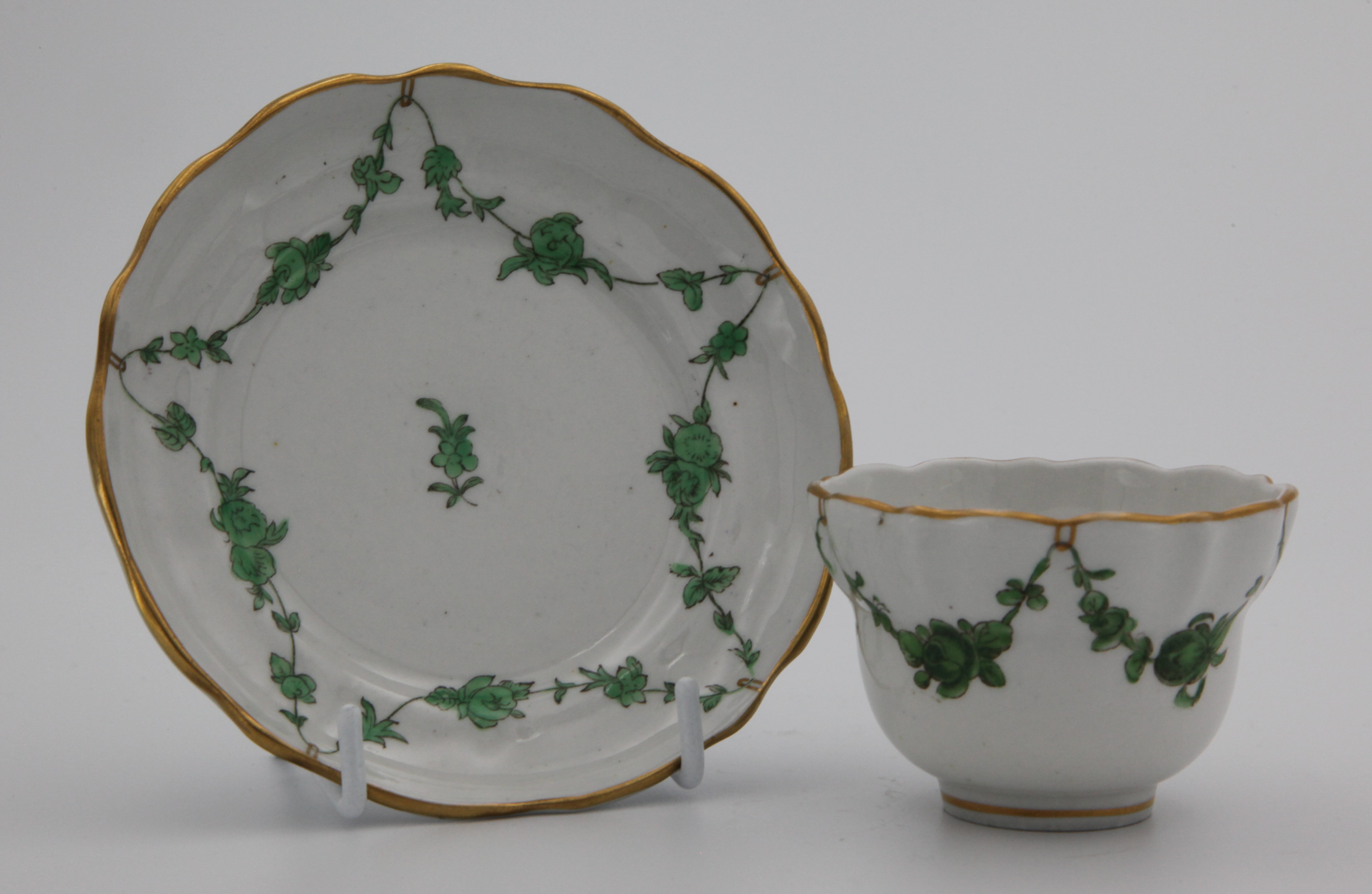 A Bristol tea bowl and saucer with green monochrome garlands suspended from gilt loops hanging from a gilt dentil rim.