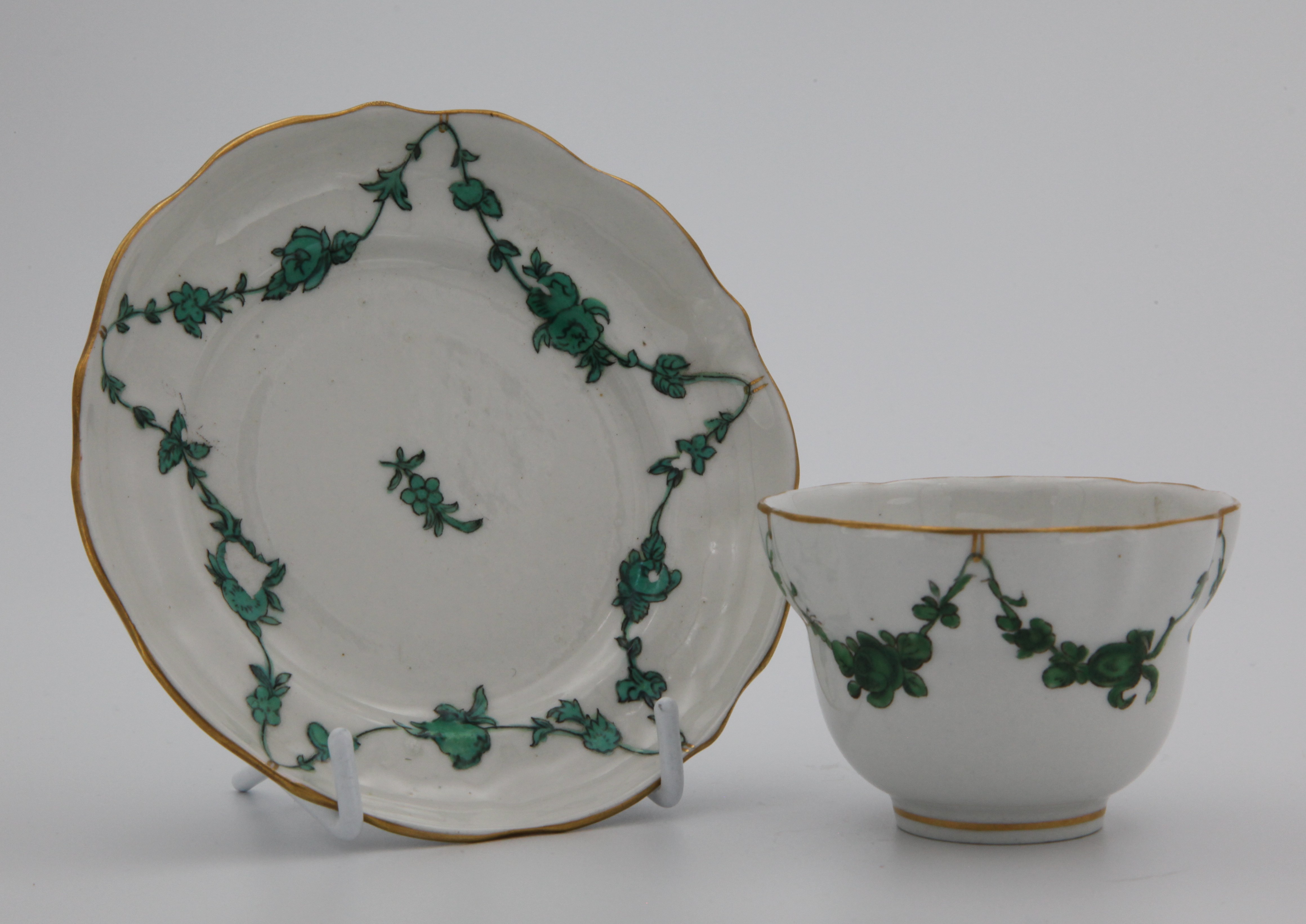 A Bristol tea bowl and saucer with green monochrome garlands suspended from gilt loops hanging from a gilt dentil rim.