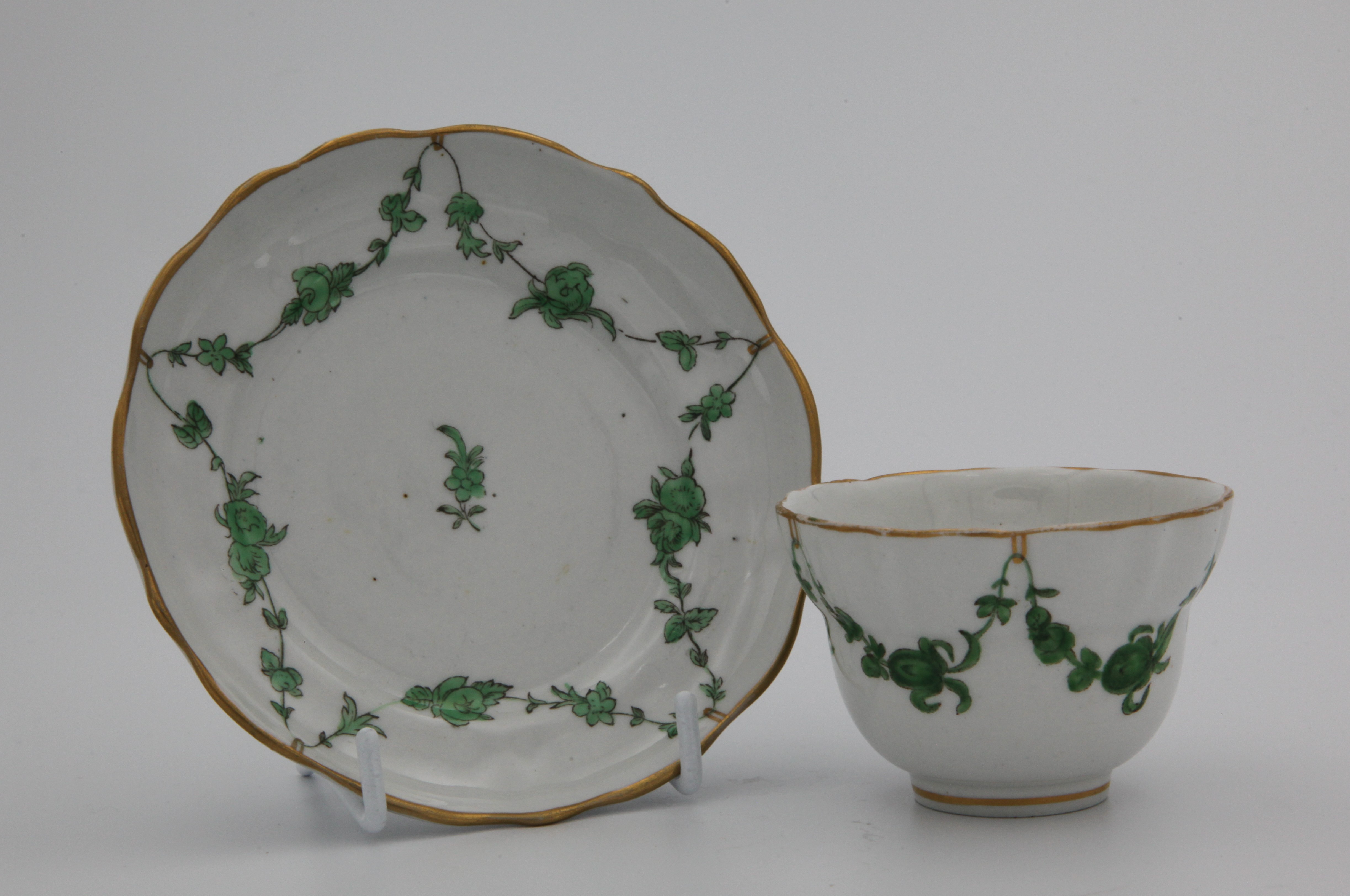 A Bristol tea bowl and saucer with green monochrome garlands suspended from gilt loops hanging from a gilt rim line.